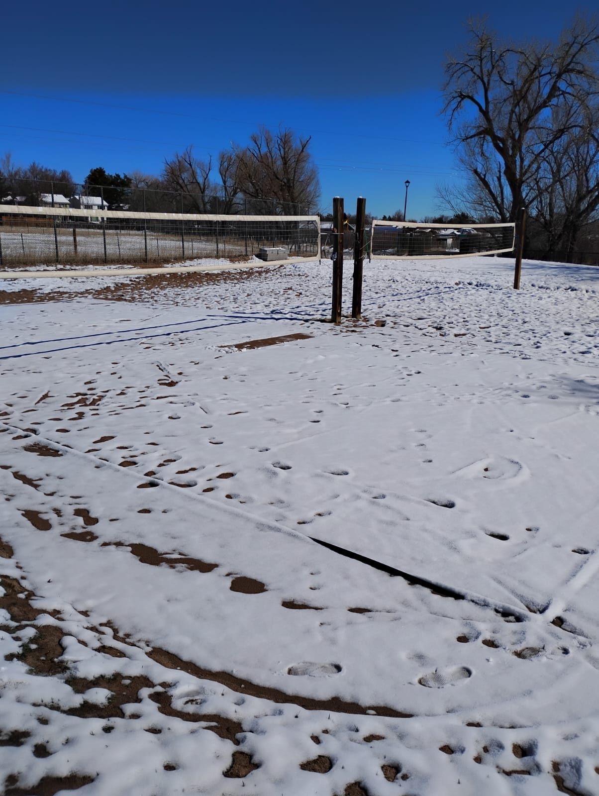The same court under a few inches of snow, footprints across it, net still up, hard blue winter sky.